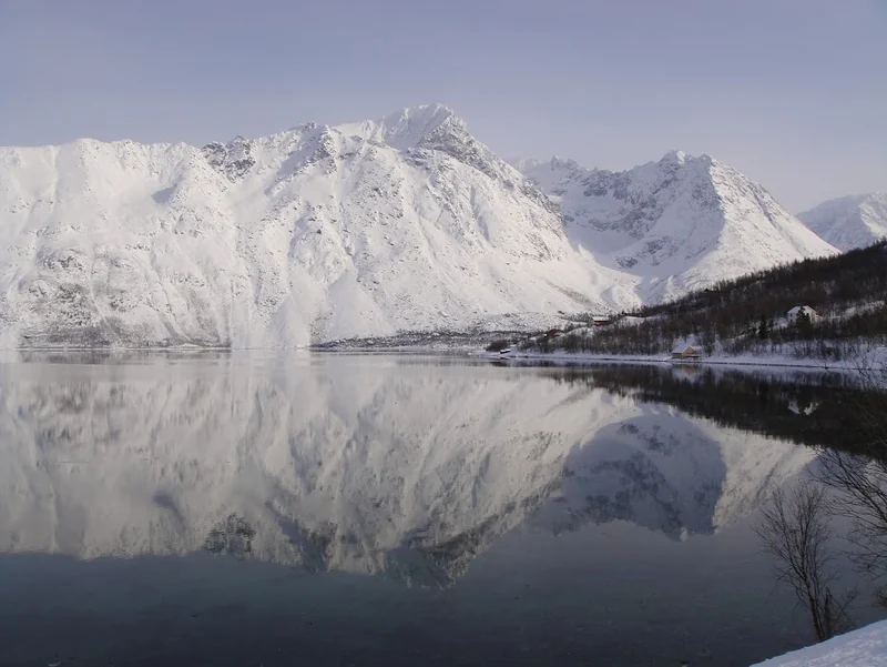 Snødekte fjell speiler seg i vann under en lys himmel